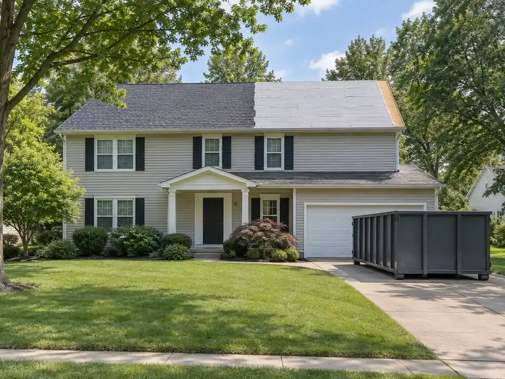 Two-story house with new shingles beside exposed underlayment and a driveway debris container.